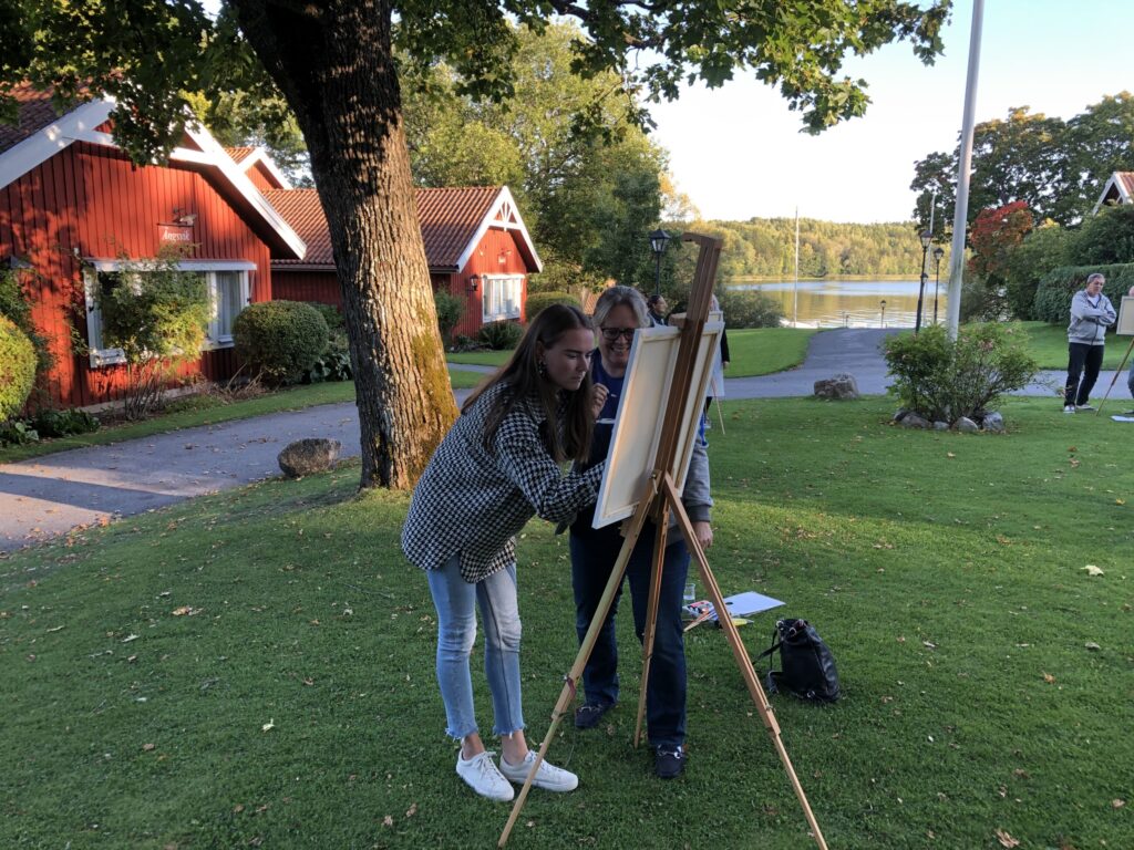 Två stycken står och målar på en tavla på ett stativ.