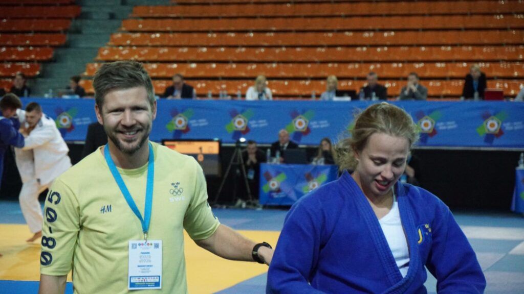 Johan Berg ler och går av tävlingsmattan tillsammans med Ida Eriksson som han coachat under en europacup i judo. I bakgrunden syns två andra som tävlar och banderoller med EJU:s logotyp. 