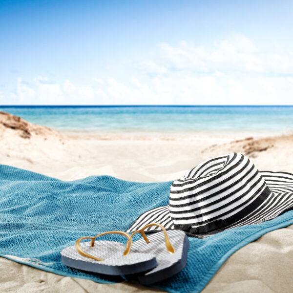 Randig hatt och flip-flops på en blå handdduk på en strand.
