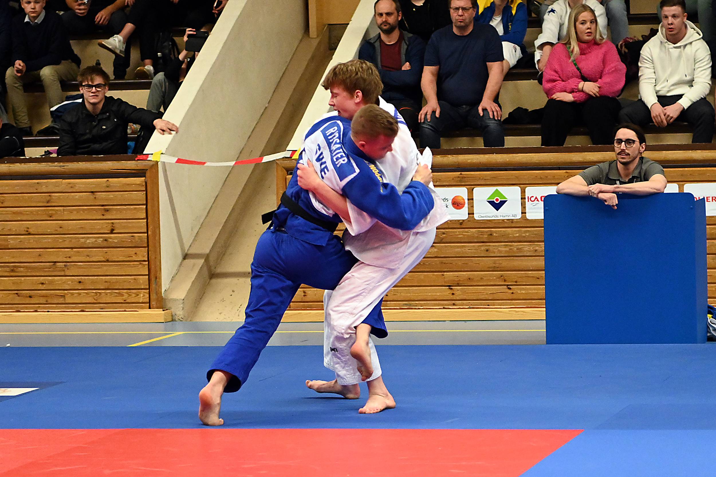 Två killar i judodräkt står på judomattan. Den ena har krokat sitt ben runt den andra och försöker fälla honom.